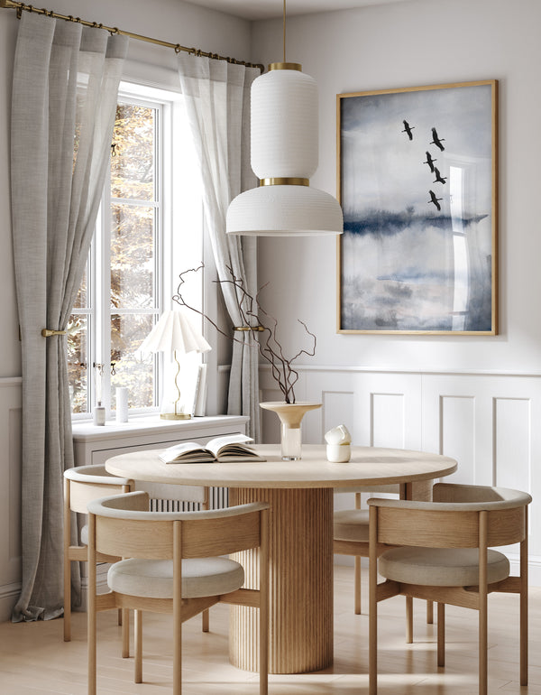A framed elegant blue and gray abstract landscape painting with a flock of black cranes flying across the sky hangs above a round dining table in a minimalist dining room.