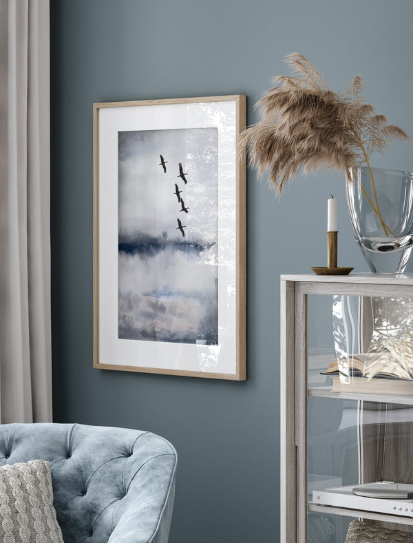 A framed elegant blue and gray abstract landscape painting with a flock of black cranes flying across the sky hangs on a wall in a living room with a deep blue accent wall.