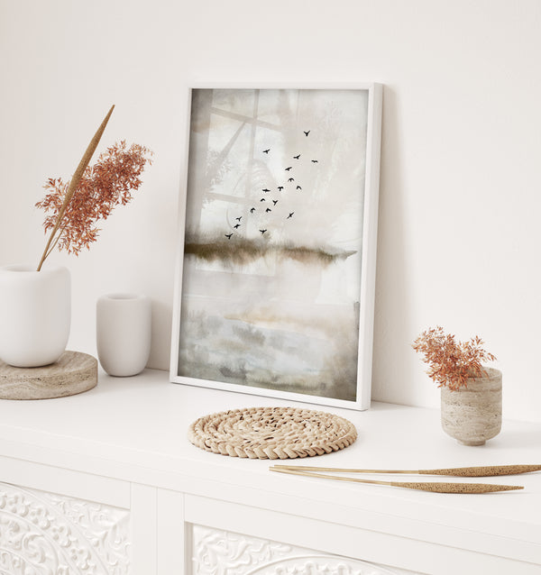 A framed watercolor painting depicting a flock of birds flying over a misty landscape. The painting is displayed on a white console table, adorned with small vases, dried flowers, and a woven coaster. The overall aesthetic is minimalist and modern, with a focus on natural elements and a sense of calm and tranquility.