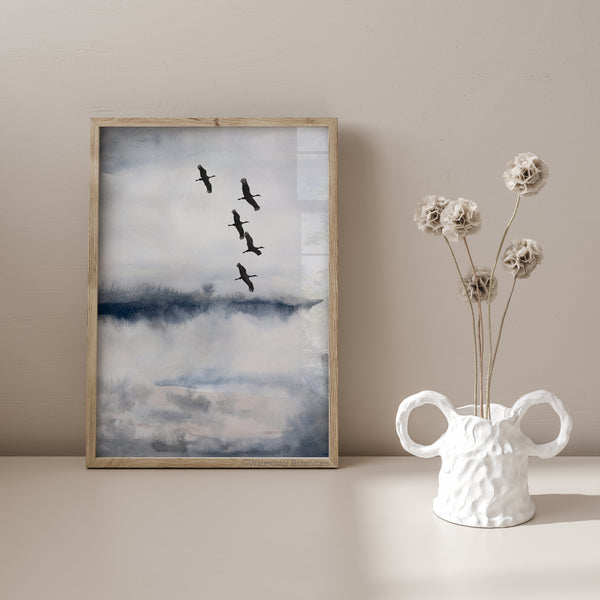 A framed elegant blue and gray abstract landscape painting with a flock of black cranes flying across the sky hangs on a wall above a small table with a vase of dried flowers.