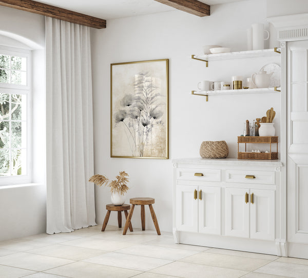 A framed print hangs on a white wall in a bright kitchen, the print features black poppies with a feathery branch, rendered in a watercolor style against a textured, off-white background, and is framed in a thin gold frame. To the left, a large window with white curtains allows natural light into the room. A small wooden stool with dried flowers in a vase sits near the window. To the right, a white kitchen cabinet with gold handles and a white countertop is visible.