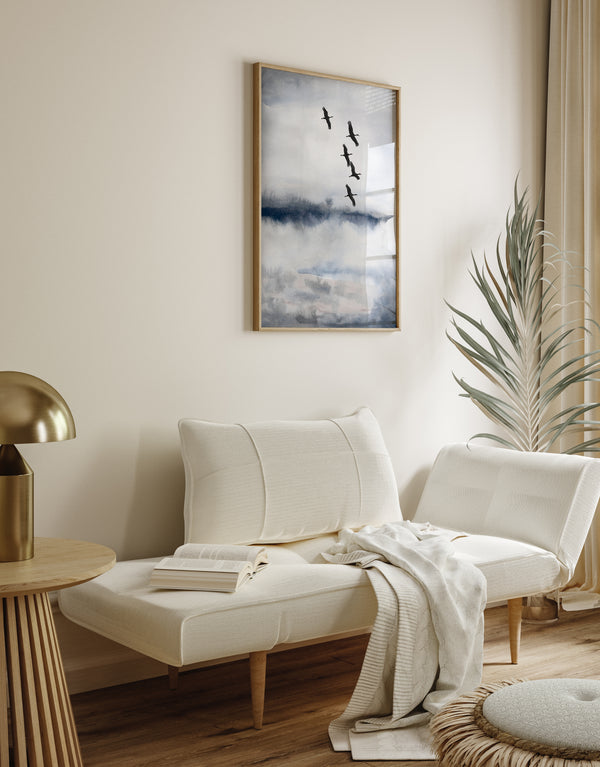 A framed elegant blue and gray abstract landscape painting with a flock of black cranes flying across the sky hangs above a cozy daybed in a minimalist living room.