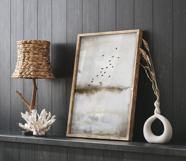 A framed watercolor painting depicting a flock of birds flying over a misty landscape. The painting is displayed on a dark wooden wall, adding a touch of warmth to the space. The painting is accompanied by a table lamp with a woven shade, a decorative coral, and a vase with dried flowers, creating a cozy and inviting atmosphere. The overall style is rustic and eclectic, with a focus on natural elements and a sense of warmth.