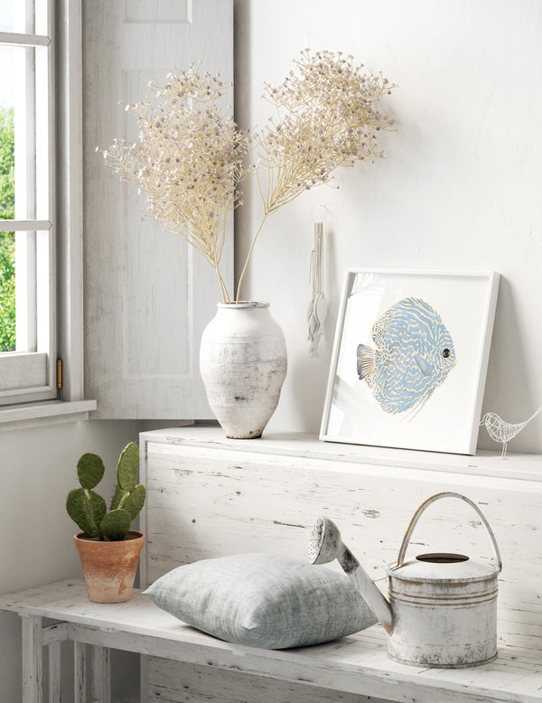 A cozy corner with a vintage feel. A framed watercolor print of a discus fish hangs on the wall above a rustic wooden bench. A vase filled with dried flowers, a small cactus in a terracotta pot, and a watering can add to the charming, rustic decor.