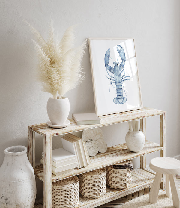 A framed watercolor painting showcases the intricate details of a blue lobster, its textured shell and powerful claws rendered with delicate precision, creating a captivating display in a coastal-themed living room with a rustic console table, dried pampas grass, and wicker baskets.