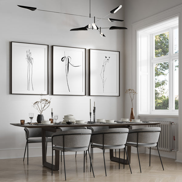 A set of three framed line drawings of various plants, rendered in sleek black lines against a crisp white background. These elegant botanical designs are displayed on a white wall above a dining table set with chairs and place settings in a modern dining room. The overall aesthetic is minimalist and sophisticated, creating a sense of calm and serenity.