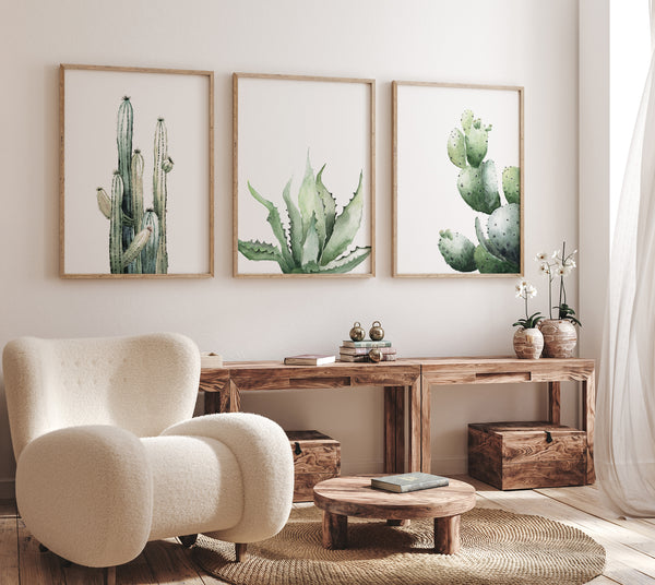 A set of three framed watercolor paintings showcasing a tall cactus, an aloe vera plant, and an opuntia cactus, displayed on a white wall in a living room setting. The paintings are framed in natural wood frames, adding a touch of warmth and texture. The living room features a comfortable armchair, a wooden coffee table, and natural light streaming in, creating a relaxing and inviting atmosphere. The cactus artwork adds a touch of the desert to this modern and stylish space.