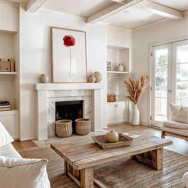 A large-scale framed watercolor painting of a single red poppy flower, displayed prominently above a marble fireplace in a spacious living room. The room features a neutral color palette with white walls, a wooden ceiling, and a cozy seating area with a plush sofa and a wooden coffee table. The overall aesthetic is warm and inviting, with a touch of rustic charm.