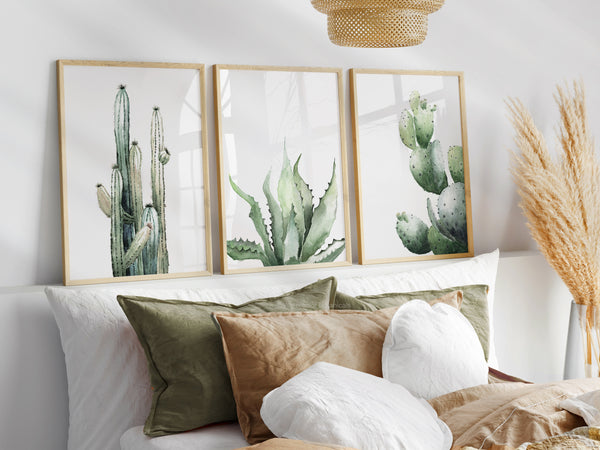 A set of three framed watercolor paintings showcasing a tall cactus, an aloe vera plant, and an opuntia cactus, displayed above a bed in a bedroom setting. The paintings are framed in natural wood frames, complementing the warm and inviting ambiance of the room. The overall aesthetic is bohemian and relaxed, with the cactus artwork adding a touch of natural beauty and a sense of adventure.