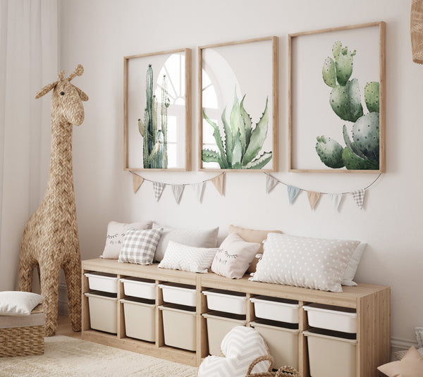 A set of three framed watercolor paintings showcasing a tall cactus, an aloe vera plant, and an opuntia cactus, displayed on a white wall in a child's playroom. The paintings are framed in natural wood frames, adding a touch of warmth and texture. The playroom features a colorful rug, a toy giraffe, and storage bins, creating a fun and playful atmosphere for a child. The cactus artwork adds a touch of whimsy and adventure to this playful space.