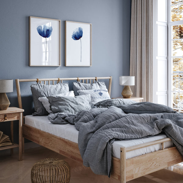 A pair of framed watercolor paintings depicting elegant navy blue poppies, displayed on a blue-toned wall above a wooden bed with a plush duvet and pillows. The room features a cozy and inviting atmosphere with natural light streaming in from a large window. The blue tones of the paintings and the bedroom decor create a sense of calm and relaxation.