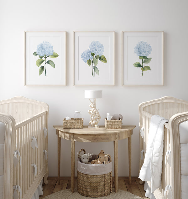 Three framed watercolor paintings of blue hydrangea flowers hang on a white wall above two cribs in a nursery.