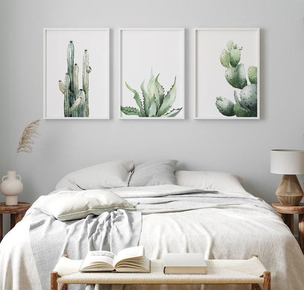 A set of three framed watercolor paintings showcasing a tall cactus, an aloe vera plant, and an opuntia cactus, displayed on a white wall in a bright and airy bedroom. The paintings are framed in natural wood frames, adding a touch of warmth and texture. The overall aesthetic is minimalist and modern, with the cactus artwork bringing a touch of the desert indoors.