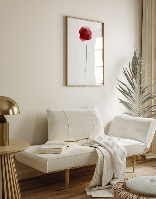 A framed watercolor painting of a single red poppy flower, displayed on a white wall above a cozy daybed with pillows and a blanket. The painting adds a vibrant pop of color to the room, creating a warm and inviting atmosphere. The overall aesthetic is minimalist and modern with a touch of bohemian flair.