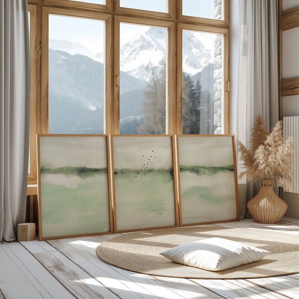 Watercolor paintings of abstract landscapes in soft sage green tones add a touch of nature to a room with a wood floor, a round rug, and a large window with a mountain view.