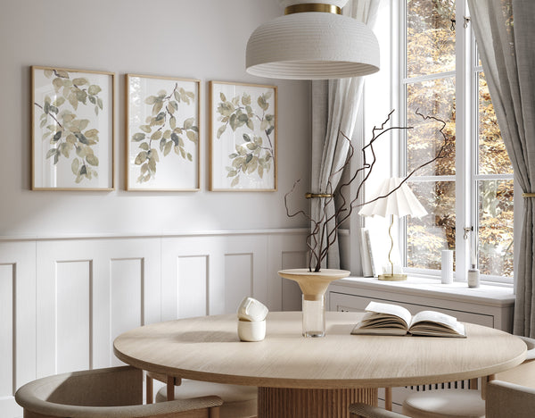 Three framed watercolor paintings of leaves in soft green hues hang on a wall above a round dining table. The room features a large window with autumnal views.