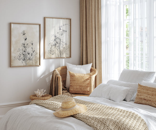 A pair of framed botanical prints featuring delicate black roses and other flowers in shades of grey and white hang above a cozy bedroom featuring a large bed dressed in white linens and a woven throw blanket.