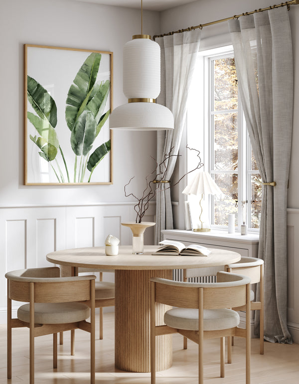 A framed watercolor painting of large tropical green banana leaves, with soft, flowing lines, hangs above a round dining table in a room with white walls and gray curtains.