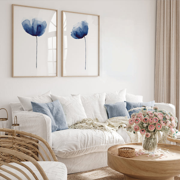 A pair of framed watercolor paintings depicting elegant navy blue poppies, displayed on a white wall above a plush white sofa with blue and white pillows. The room is filled with natural light, creating a serene and inviting atmosphere. The overall design is minimalist and modern with a touch of bohemian flair, with the poppy prints adding a touch of color and visual interest.