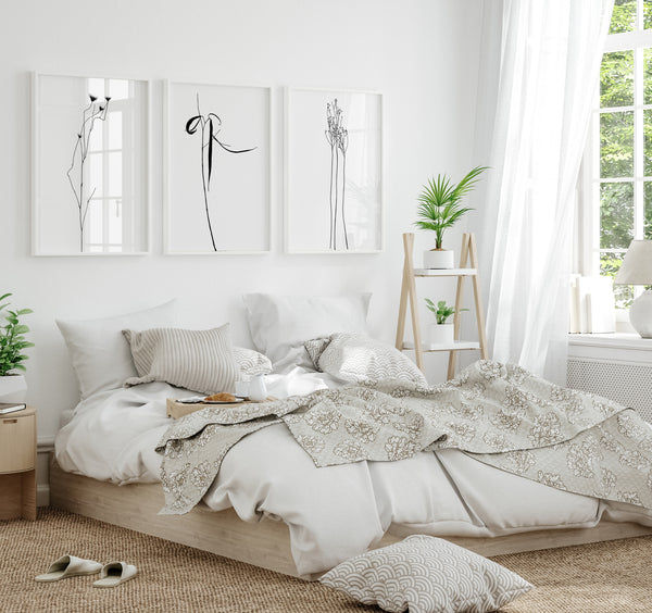 A set of three framed line drawings of various plants, rendered in sleek black lines against a crisp white background. These elegant botanical designs are displayed on a white wall above a plush bed with a neutral-colored linen bedding set in a light and airy bedroom. The overall aesthetic is minimalist and modern, creating a sense of calm and serenity.