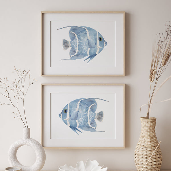 Two watercolor paintings of blue angelfish, each framed in a natural wood frame. The fish are facing each other in a horizontal orientation, creating a sense of balance and symmetry. The paintings are displayed on a white wall with dried flowers and decorative vases adding a touch of natural beauty to the space. The overall setting suggests a coastal or beach-inspired theme.