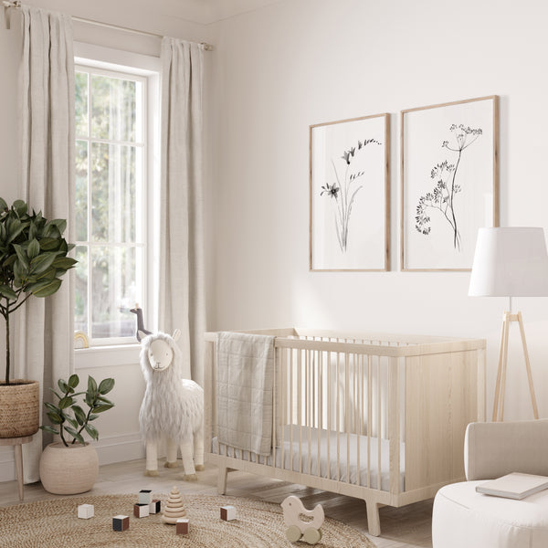 A light natural wood baby crib in a bright, modern nursery with beige walls, neutral curtains, a white sheep toy, and two framed botanical prints on the wall.