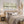 A styled bedroom scene featuring a bed with neutral-toned bedding, a woven rattan headboard and footboard, two bedside tables with lamps, three framed abstract landscape prints on the wall, and dried pampas grass decor.