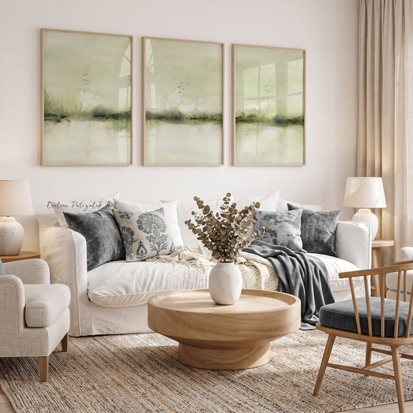 A styled living room featuring a three-panel framed wall art set with soft sage green and beige Japandi abstract landscape designs, hung above a white sofa with gray and blue patterned pillows.