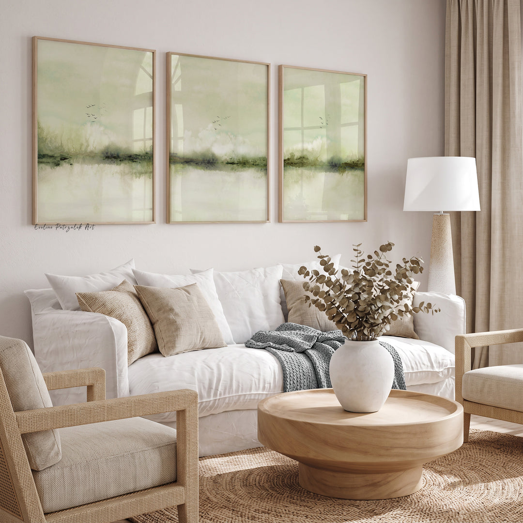 A styled living room featuring a three-panel framed wall art set with soft sage green abstract landscape prints hanging above a white sofa with neutral-toned pillows and a throw blanket.