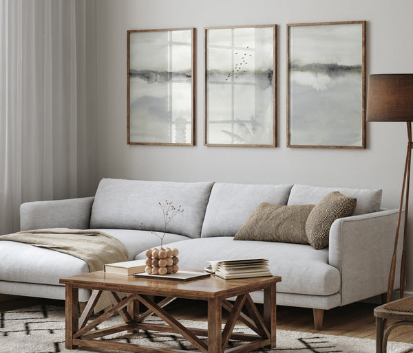 Three rectangular, medium-toned wood-framed prints featuring abstract landscape scenes in muted shades of gray and white are displayed above a light gray sofa in a living room. The prints feature soft, blended watercolor-like textures with a suggestion of a horizon line and faint, silhouetted birds in the middle print. The sofa has several textured pillows in shades of brown and gray. A dark wood coffee table with a geometric design sits in front of the sofa, holding books and a decorative object.