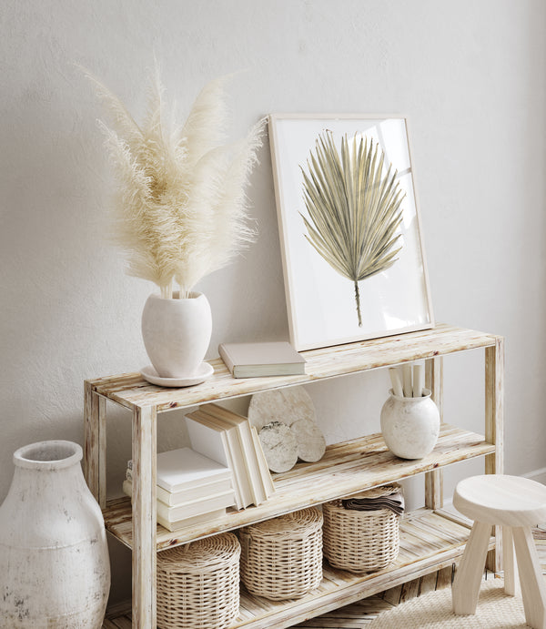 A large-scale botanical print of a palmetto leaf, framed in a natural wood frame, hangs on a white wall above a wooden console table. The table is styled with a vase of dried flowers, a stack of books, and decorative objects, including a vase, a lamp, and a string of beads. The overall style is coastal and minimalist with a focus on natural elements and a relaxed, beachy vibe. The presence of a vase of dried pampas grass further enhances the natural and bohemian aesthetic of the space.