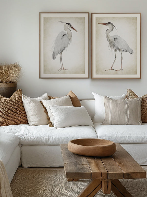 A pair of framed watercolor paintings depicting elegant great blue herons in soft neutral tones against a vintage-inspired background. The paintings are displayed on a white wall above a comfortable sofa in a living room with a natural and relaxed atmosphere.