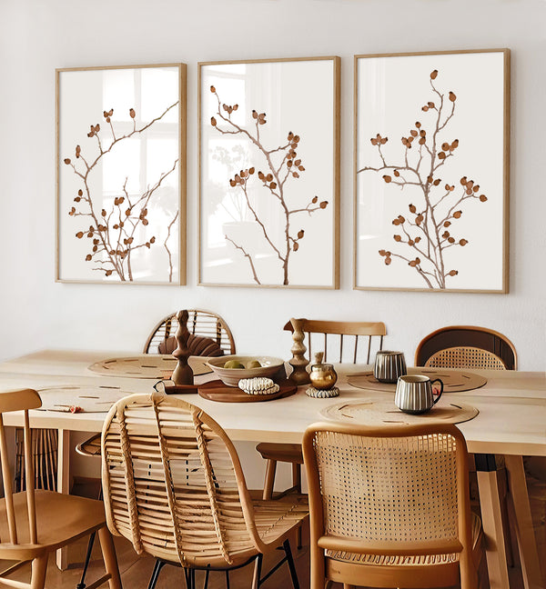 Three framed watercolor paintings showcase the intricate details of rose hip branches, painted in rich burnt sienna tones against a crisp white background, creating a stunning display above a dining table with a rustic wooden top and woven chairs in a living room.