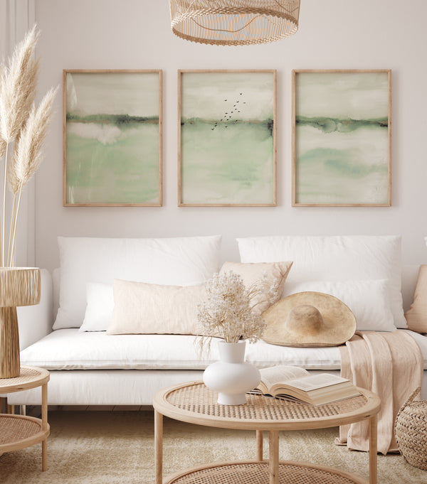 Watercolor paintings of abstract landscapes in soft sage green tones add a touch of nature to a living room with a white sofa, a round coffee table, and a wicker hanging lamp.