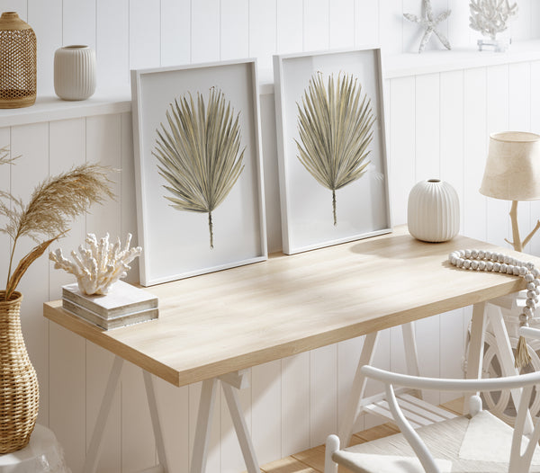 Two large-scale botanical prints of palmetto leaves, each framed in a white frame, hang on a white wall above a wooden desk. The desk is adorned with a variety of decorative objects, including a vase, a lamp, and a string of beads. The overall style is coastal and minimalist with a focus on natural elements and a relaxed, beachy vibe.