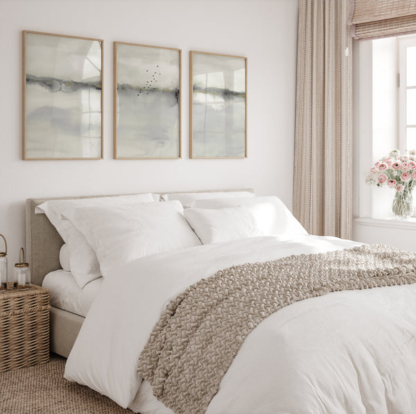 Three rectangular, light wood-framed prints featuring abstract landscape scenes in muted shades of gray and white are displayed above a queen-sized bed with white linens. The prints feature soft, blended watercolor-like textures with a suggestion of a horizon line and faint, silhouetted birds in the middle print. A beige, textured throw blanket is folded at the foot of the bed. On the left is a round wicker side table with a glass container and a small lamp. A window with striped curtains and a vase of pink