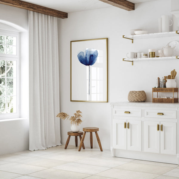 A framed watercolor painting depicting an elegant navy blue poppy, displayed on a white wall in a kitchen setting. The painting is framed in a simple, natural wood frame, complementing the clean lines of the kitchen cabinetry. The kitchen features a large window with white curtains, white cabinets, and a wooden countertop, creating a bright and airy atmosphere. The overall design is minimalist and modern with a touch of bohemian flair, with the poppy print adding a touch of color and visual interest.