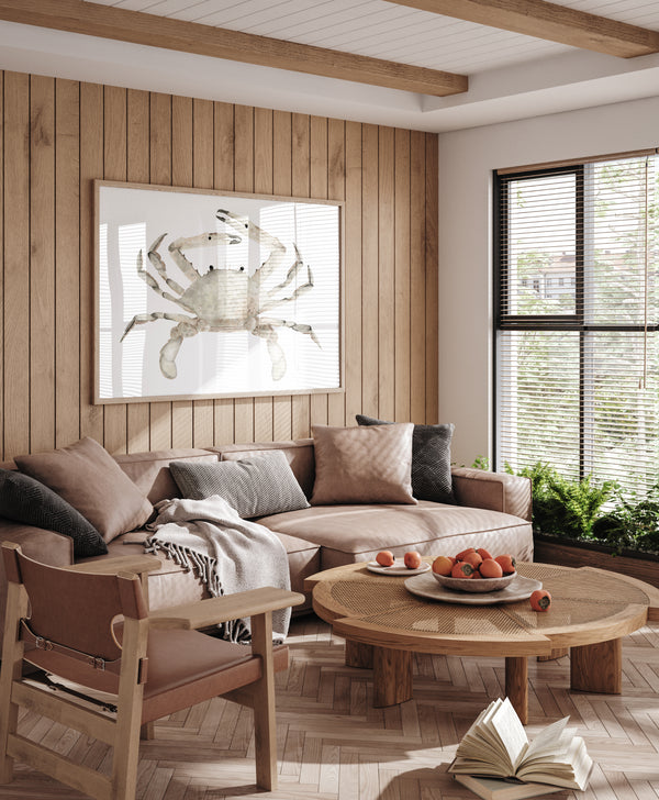 A large, framed watercolor painting of a realistic crab in soft, earthy brown tones hangs prominently on a wood-paneled wall in a cozy living room. The painting adds a touch of coastal charm to the space, which is filled with comfortable seating, a coffee table, and natural light streaming in from a large window.