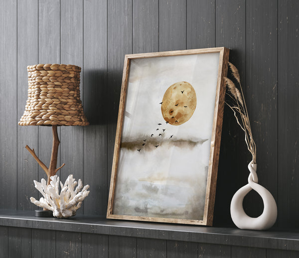 A watercolor painting depicting a large, full moon with a muted yellow hue. A flock of black birds flies across the moon's face, creating a dramatic contrast. The background is a soft, muted grey with subtle hints of blue and green. The painting is displayed on a dark wooden shelf, with a wicker lampshade and a decorative coral piece on either side.