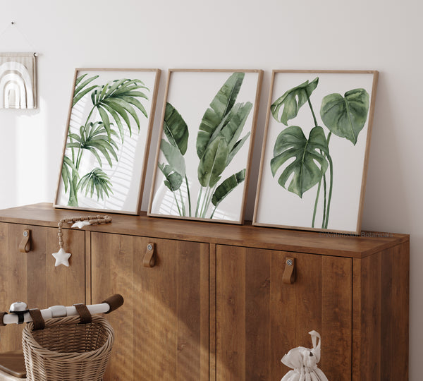 Three rectangular, light wood-framed prints featuring detailed watercolor illustrations of various tropical plants with large green leaves are displayed above a dark wood cabinet with two doors and silver pulls. On the cabinet to the left is a light wood and white beaded garland with a star and tassel detail. A portion of a brown wicker basket is visible in the bottom left corner, and a small white figurine is visible in the bottom right. The wall behind the cabinet is white.