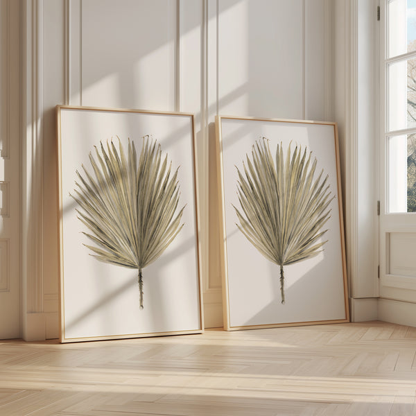 Two large-scale botanical prints of palmetto leaves, each framed in a natural wood frame. The leaves are depicted in a detailed watercolor style with variations in shades of green and brown, creating a realistic and vibrant effect. The prints are displayed on a white wall in a spacious room with hardwood floors and large windows, creating a bright and airy atmosphere. The overall mood of the image is serene and tranquil, evoking a sense of connection to nature.