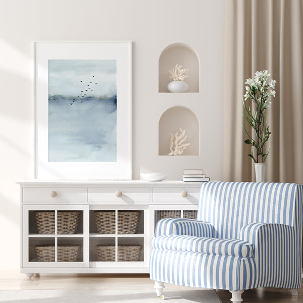 A framed Blue Watercolor Abstract painting of a blue and white landscape with a flock of birds flying across the sky hangs above a white dresser in a coastal-inspired living room.