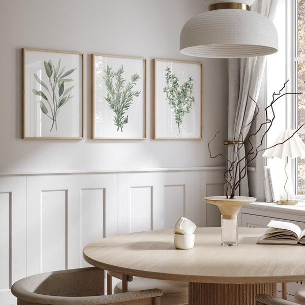 Three framed watercolor paintings showcase delicate depictions of Provencal herbs, including sage, rosemary, and thyme, in soft muted green tones, creating a calming and inviting atmosphere above a dining table with a round wooden top and woven chairs."