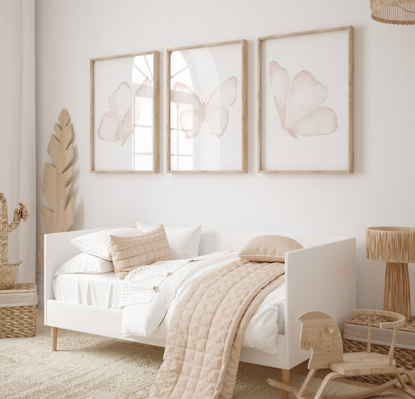 A set of three framed watercolor paintings of delicate pink butterflies, arranged in a row on a light-colored wall above a daybed in a child's bedroom. The butterflies are depicted with soft, ethereal details, creating a sense of lightness and airiness. The room is filled with soft textures and natural light, creating a serene and inviting space for a child. The overall aesthetic is whimsical and playful, with the butterfly art adding a touch of magic and wonder.