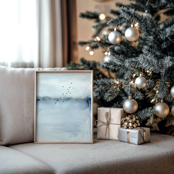 A framed Blue Watercolor Abstract painting of a blue and white landscape with a flock of birds flying across the sky sits on a sofa next to a decorated Christmas tree.