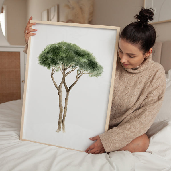 A woman lovingly cradles a framed watercolor painting. The painting showcases a pine tree with a lush, rounded canopy and two intertwined trunks, rendered in delicate shades of green against a crisp white background.