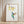 A modern hummingbird fine art poster leans against a white paneled wall with decorative molding on a light wood herringbone floor. The poster features a watercolor illustration of a hummingbird with blue, green, and brown tones, its wings spread, positioned below a branch with yellow, bell-shaped flowers. The poster is framed in a thin, light wood frame.