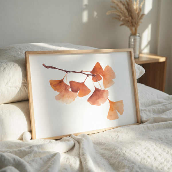 A framed watercolor art print of ginkgo leaves in warm orange and brown tones, leaning against a bed with white textured bedding in a softly lit bedroom.