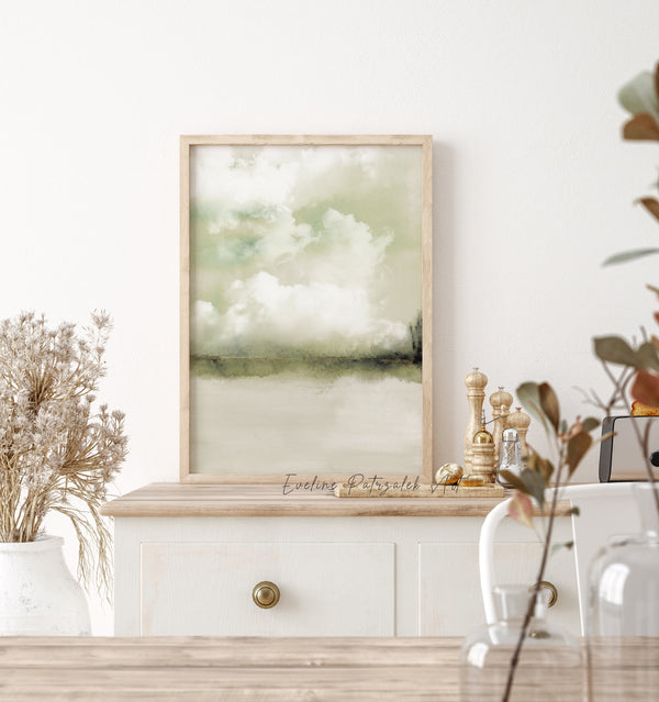 A framed watercolor landscape print featuring soft green and white clouds over a calm lake, styled on a white wooden cabinet in a bright, modern room.