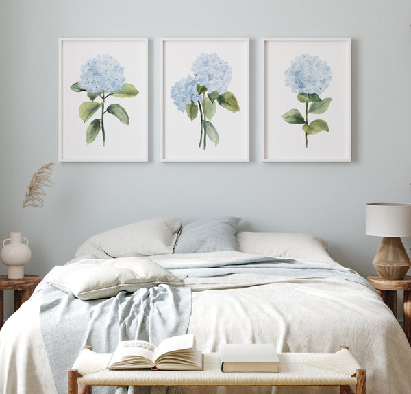 Three framed watercolor paintings of blue hydrangea flowers with green leaves and stems adorn a light blue wall above a bed with white linens in  a bedroom. A wooden nightstand with a lamp and a book completes the scene.